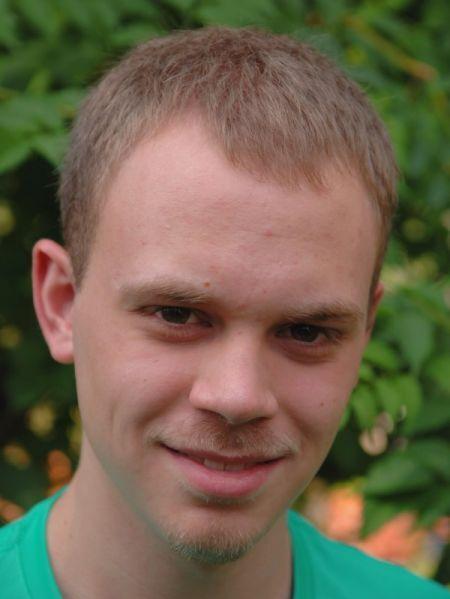 Portr&auml;t eines jungen Mannes mit kurzem Haar und einem l&auml;chelnden Gesichtsausdruck vor gr&uuml;nem Hintergrund.