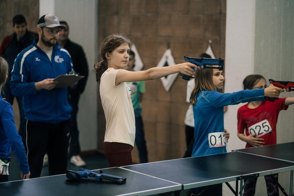 M&auml;dchen zielen mit Luftgewehren auf eine Zielscheibe, w&auml;hrend Erwachsene die Veranstaltung beobachten.