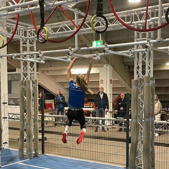 Athletin schwingt an einem Trainingsger&uuml;st mit Ringen und Balken, Menschen verfolgen die &Uuml;bungen im Hintergrund.