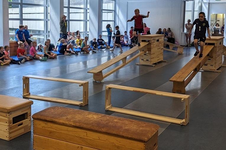 Zwei Kinder balancieren auf Holzbalken in einer Sporthalle, w&auml;hrend eine Gruppe Zuschauer zuschaut.