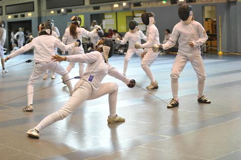 Mehrere Fechter in Schutzkleidung &uuml;ben auf einem Sportboden, einer f&uuml;hrt einen Torchken mit einem Degen aus.