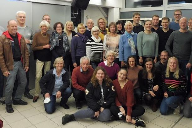 Gruppenfoto von etwa 40 Personen in einem Raum, einige stehen, andere sitzen, in verschiedenen Altersgruppen und Kleidung.