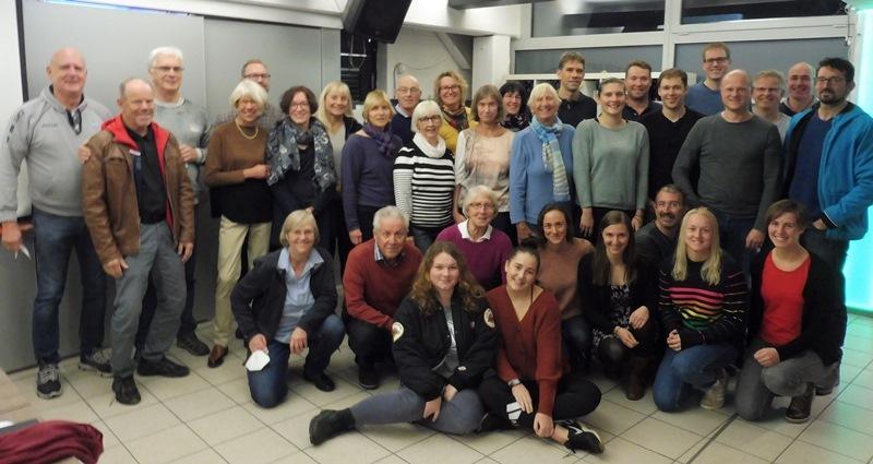 Gruppenfoto von etwa 40 Personen in einem Raum, einige stehen, andere sitzen, in verschiedenen Altersgruppen und Kleidung.