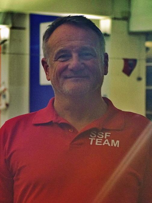 Mann mit grau melierten Haaren und freundlichem L&auml;cheln in rotem Shirt mit &bdquo;SSF TEAM&ldquo; vor einem Schwimmbecken.