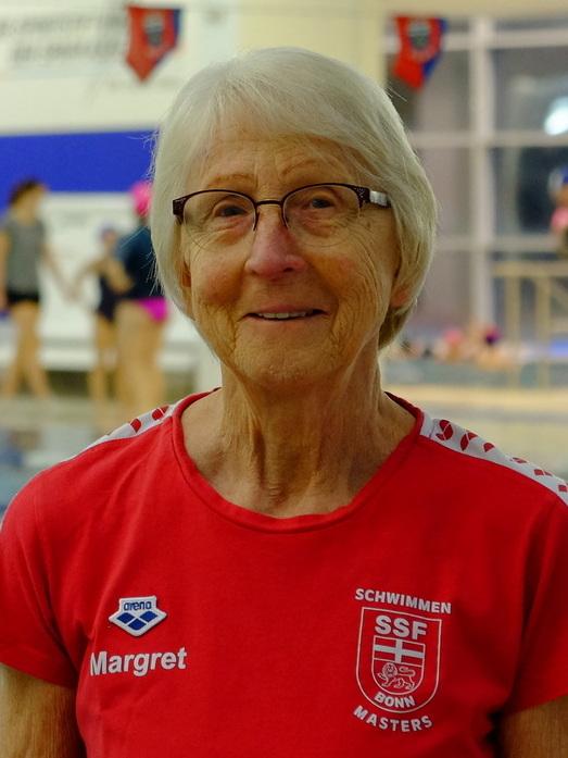 &Auml;ltere Schwimmerin mit Brille in rotem Shirt der SSF Bonn, Hintergrund mit Schwimmbecken und &Uuml;benden.