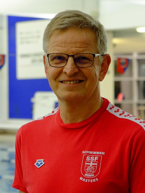 L&auml;chelnder Mann in rotem T-Shirt mit Schwimmen-Logo steht am Beckenrand eines Schwimmbads.