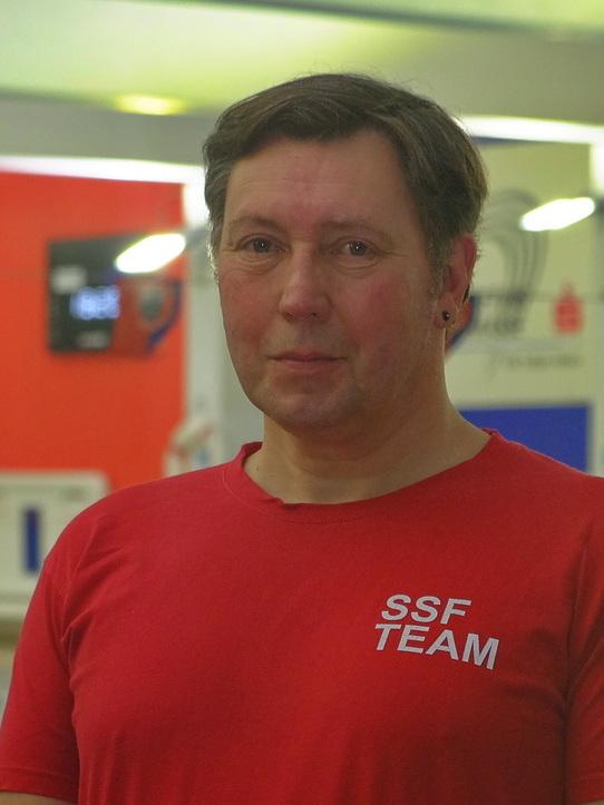 L&auml;chelnder Mann in rotem T-Shirt mit &bdquo;SSF TEAM&ldquo;-Aufdruck, stehend in einer Schwimmhalle im Hintergrund.