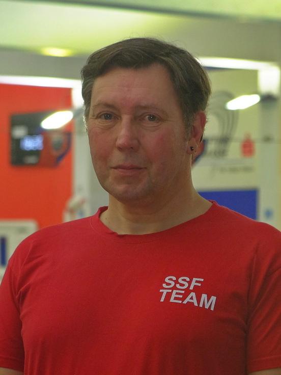 L&auml;chelnder Mann in rotem T-Shirt mit &bdquo;SSF TEAM&ldquo;-Aufdruck, stehend in einer Schwimmhalle im Hintergrund.