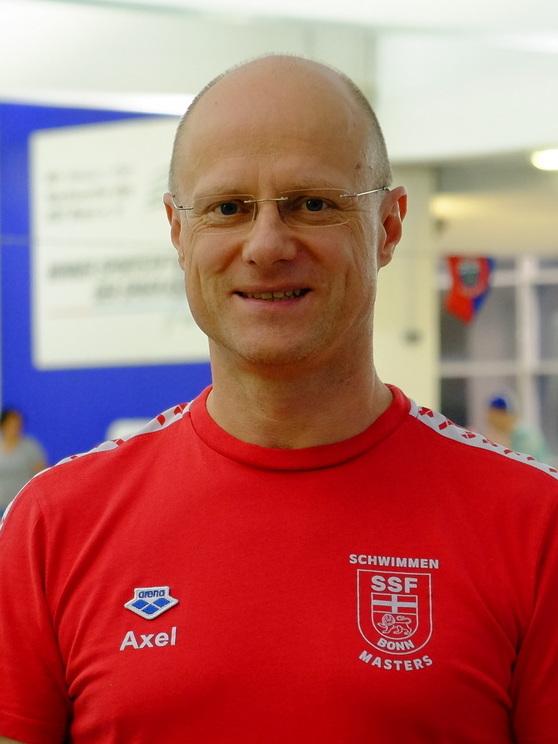 Mann mit Brille in rotem Schwimmtrikot steht am Beckenrand, Hintergrund zeigt Schwimmbecken und Sportbanner.