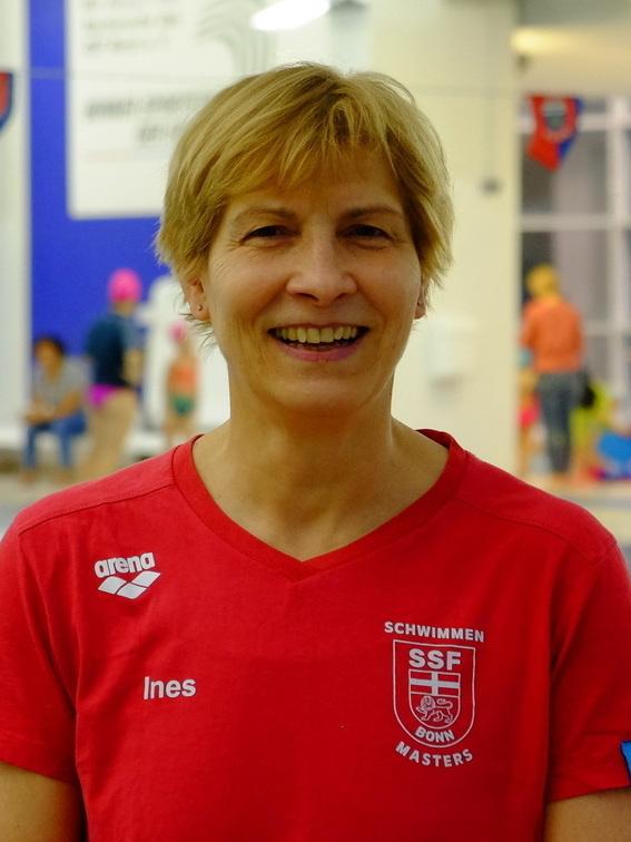 L&auml;chelnde Frau in rotem Schwimmshirt steht am Beckenrand eines Schwimmbads, im Hintergrund sind Schwimmer zu sehen.