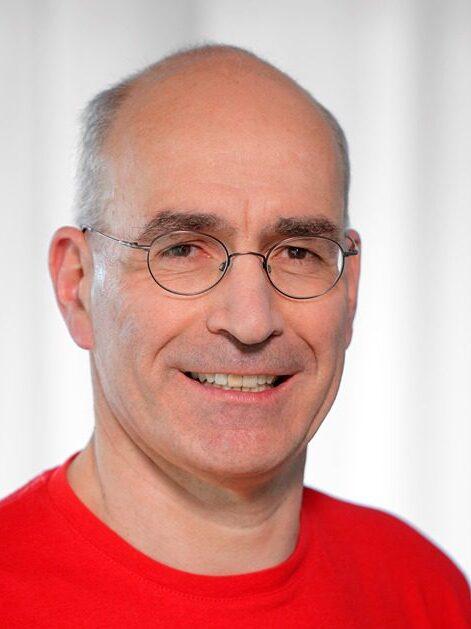 Portr&auml;t eines Mannes mit Brille, der in einem roten Shirt l&auml;chelt, vor hellem, verschwommenem Hintergrund.