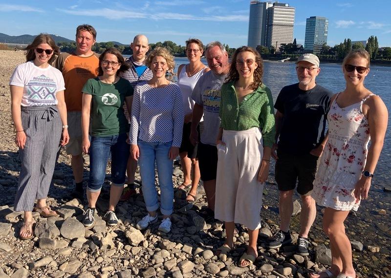 Gruppe von zehn Personen steht l&auml;chelnd am Ufer eines Flusses, hinter ihnen sind Geb&auml;ude und B&auml;ume sichtbar.