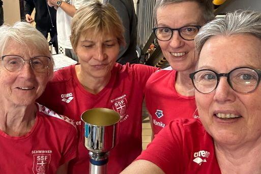 Vier Frauen in roten Shirts posieren mit einem Pokal in der Mitte, l&auml;cheln und feiern in einer Sporthalle.