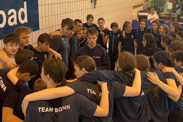 Zusammenkunft von Schwimmern in Teamkleidung, die sich umarmen und sich auf ein Event vorbereiten, im Hintergrund ein Schwimmbad.