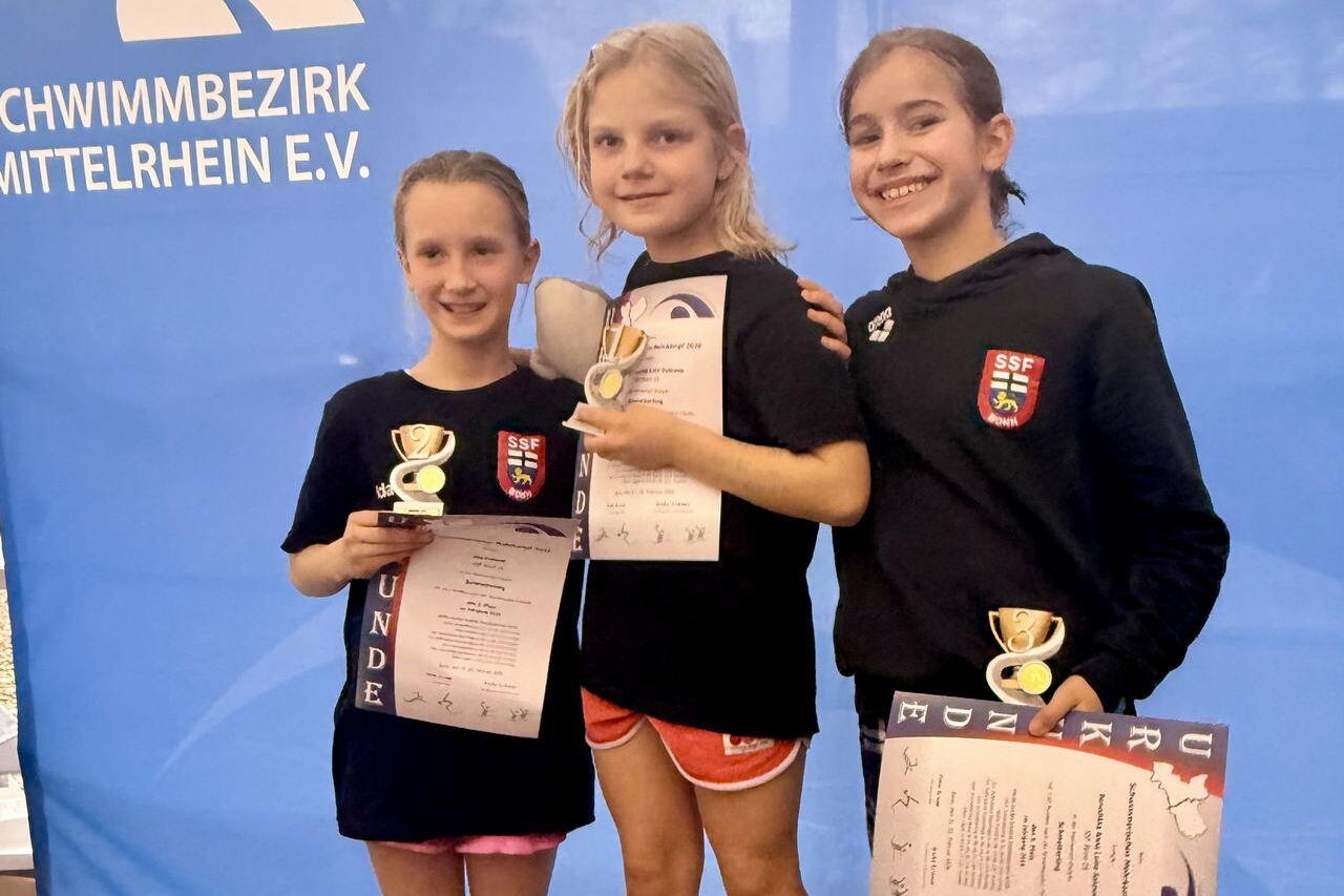 Drei M&auml;dchen stehen auf einem Podest, halten Medaillen und Urkunden, vor einem blauen Banner des Schwimmbezirks Mittelrhein.