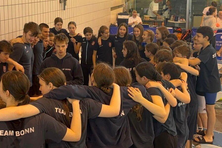 Zusammenkunft von Schwimmern in Teamkleidung, die sich umarmen und sich auf ein Event vorbereiten, im Hintergrund ein Schwimmbad.
