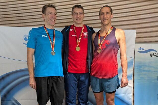 Drei Medaillengewinner stehen l&auml;chelnd auf einem Siegerpodest mit Medaillen um den Hals.
