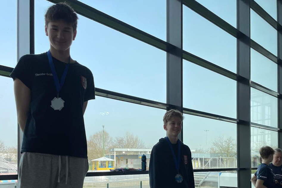 Zwei Jungen mit Medaillen stehen neben einem gro&szlig;en Fenster, im Hintergrund sind Sporttaschen und eine Wiese sichtbar.