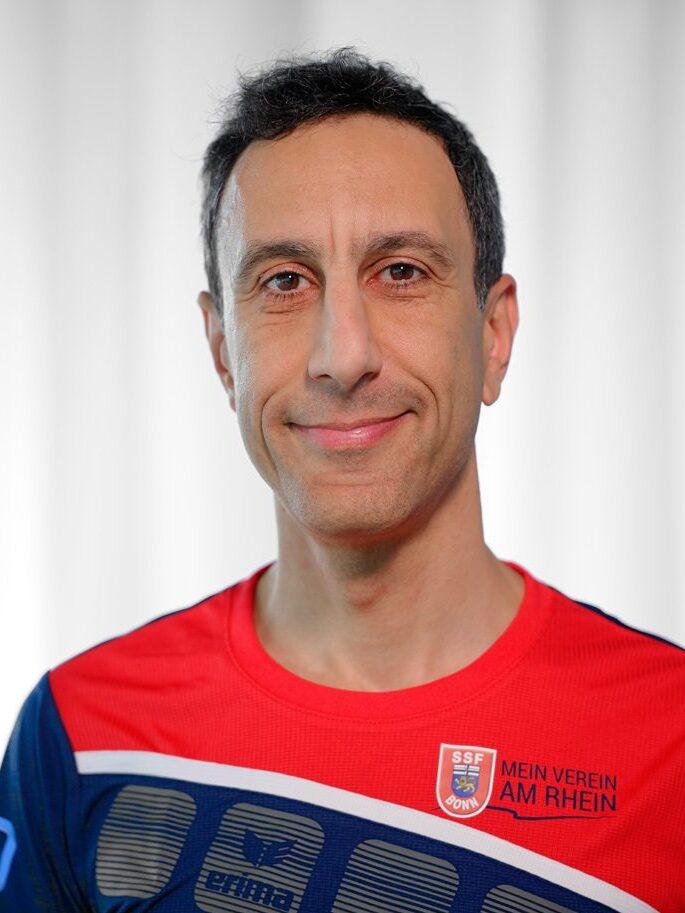 Portr&auml;t eines Mannes in einem rot-blauen Sporttrikot mit dem Schriftzug &bdquo;Mein Verein am Rhein&ldquo; vor neutralem Hintergrund.