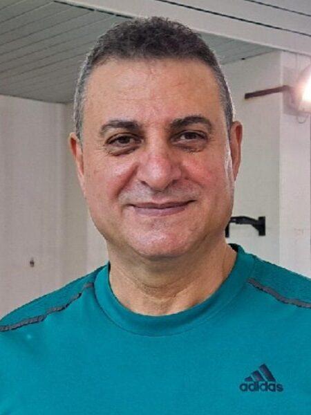 Portr&auml;t eines l&auml;chelnden Mannes in einem t&uuml;rkisfarbenen T-Shirt, im Hintergrund eine helle Sporteinrichtung.