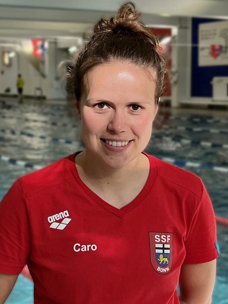 L&auml;chelnde Frau in rotem Shirt mit Aufdruck &bdquo;Caro&ldquo; steht vor einem Schwimmbecken im Hintergrund.