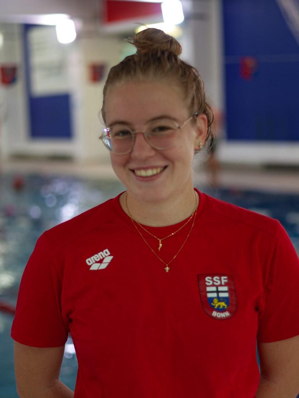 Eine junge Frau mit Brille l&auml;chelt in rotem T-Shirt am Beckenrand eines Schwimmbads. Hintergrund mit Wasser und blauen W&auml;nden.