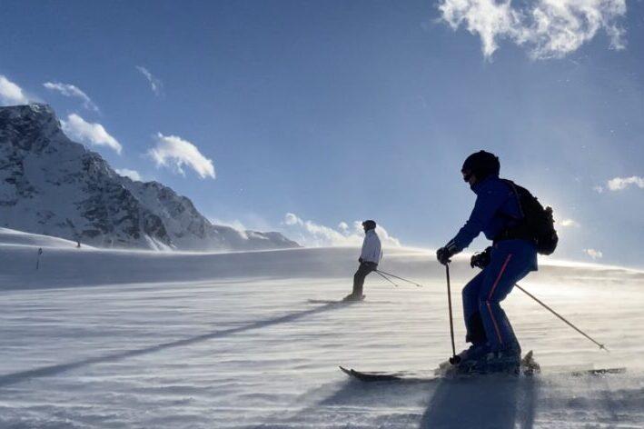 Zwei Skifahrer fahren auf einer verschneiten Piste mit Bergen und blauen Himmel im Hintergrund.