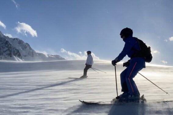 Zwei Skifahrer fahren auf einer verschneiten Piste mit Bergen und blauen Himmel im Hintergrund.