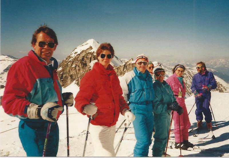 Sechs Personen in bunter Ski-Ausr&uuml;stung stehen an einem schneebedeckten Berghang mit Bergen im Hintergrund.