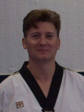 Person in schwarzer und wei&szlig;er Taekwondo-Gewandung steht vor einer hellen Wand. Freundlicher Gesichtsausdruck.