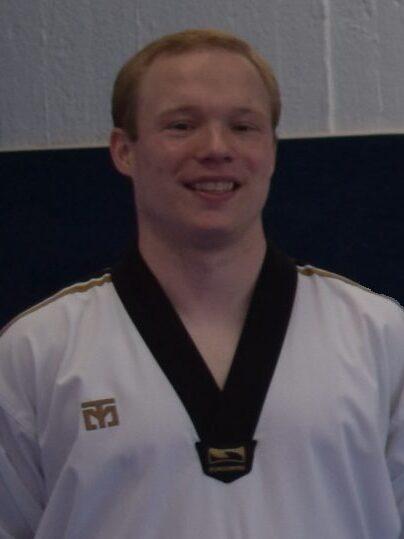 L&auml;chelnder Mann in einem wei&szlig;en Taekwondo-Gi mit schwarzem Kragen, vor einer blauen Wand.