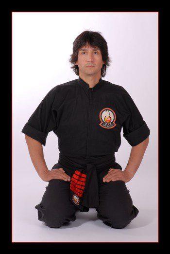 Mann im schwarzen Karateanzug sitzt kniend mit einem ruhigen Ausdruck vor neutralem Hintergrund.