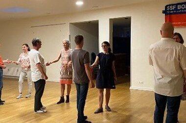 Eine Gruppe von sieben Personen tanzt gemeinsam in einem hellen Raum mit Holzfu&szlig;boden.