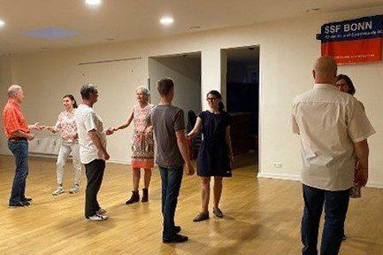 Eine Gruppe von sieben Personen tanzt gemeinsam in einem hellen Raum mit Holzfu&szlig;boden.