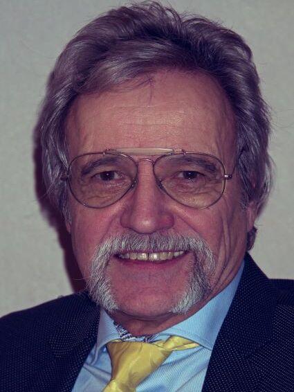 Portr&auml;t eines &auml;lteren Mannes mit grauen Haaren, Brille und Gehstock, der in die Kamera l&auml;chelt. Er tr&auml;gt einen Anzug mit Krawatte.