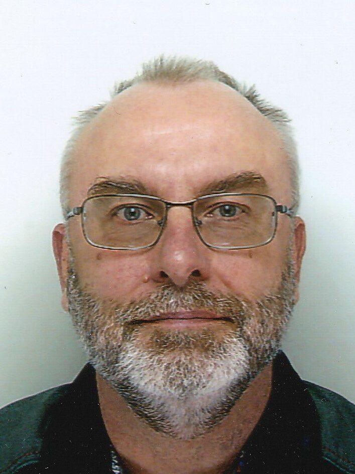 Portr&auml;t eines Mannes mit Brille, grauem Bart und Haar, vor einem neutralen Hintergrund. Er tr&auml;gt ein schwarzes Oberteil.