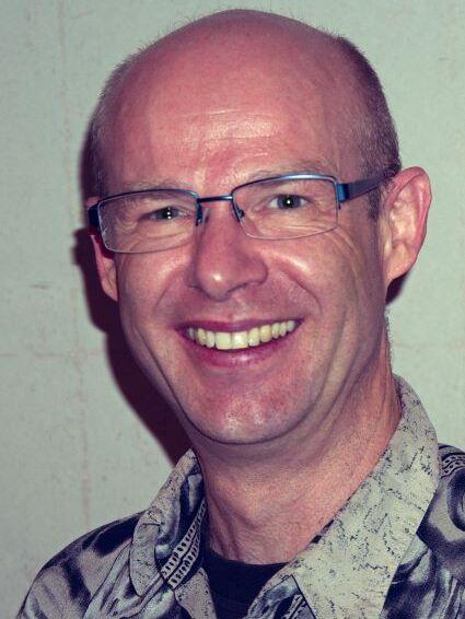 Ein l&auml;chelnder Mann mit Brille tr&auml;gt ein gemustertes Hemd, vor einem hellen Hintergrund.