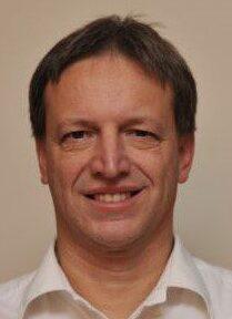 Nahaufnahme eines Mannes mit kurzem, grauem Haar und L&auml;cheln, bekleidet mit einem wei&szlig;en Hemd.