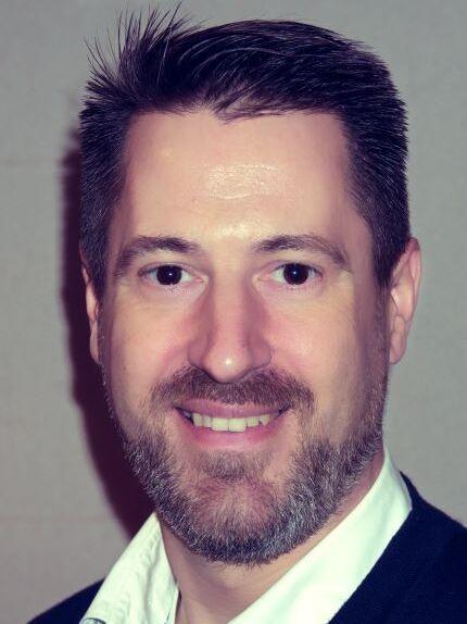 Portr&auml;t eines Mannes mit Bart, kurzes Haar und l&auml;chelndem Gesicht vor neutralem Hintergrund.