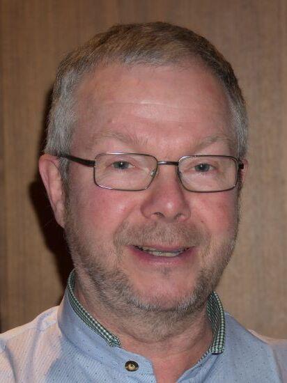 Nahaufnahme eines l&auml;chelnden Mannes mit Brille, hellblauem Hemd und grauen Haaren vor einer Holzpaneele.