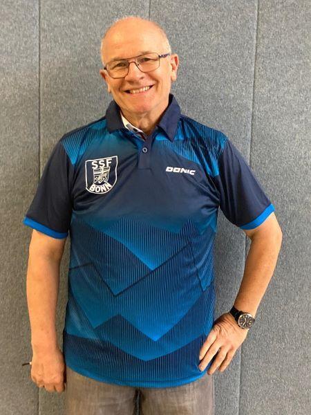 Mann in blauer Sportkleidung mit Logo von SSF Bonn l&auml;chelt vor einer grauen Wand.