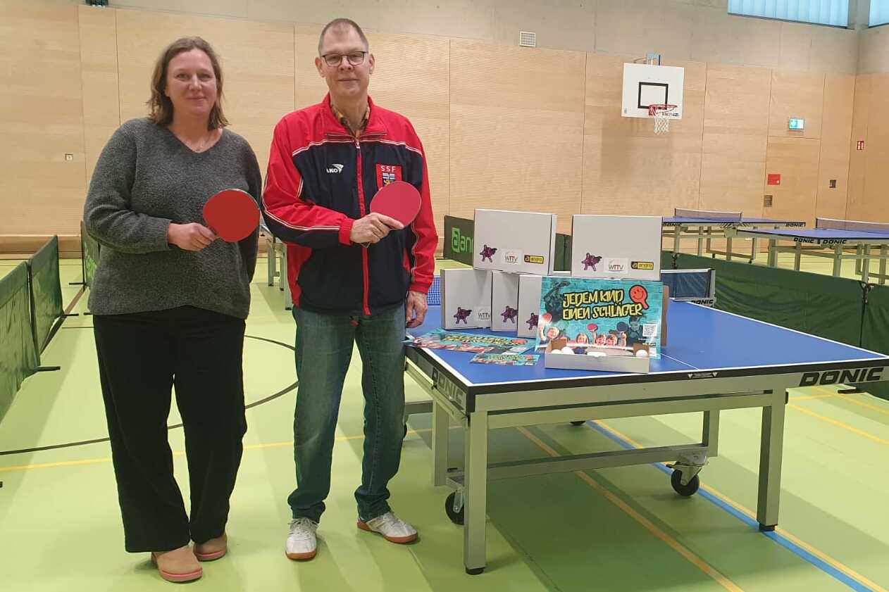 Zwei Personen mit Tischtennisschl&auml;gern stehen neben einem Tischtennistisch, umgeben von Spielger&auml;ten und -materialien.