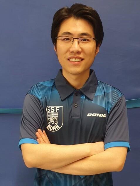 Junger Mann mit kurzer Haarfrisur tr&auml;gt ein blaues Tischtennis-Trikot und steht vor einem blauen Hintergrund.