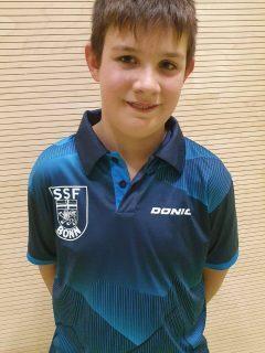 Junge mit Kurzhaarschnitt tr&auml;gt ein blaues Poloshirt mit Vereinslogo und steht vor einer hellen Wand.