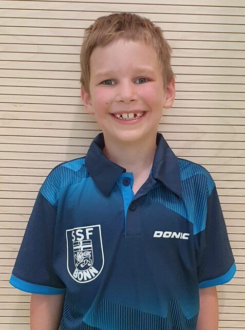 L&auml;chelnder Junge in blauem Tischtennis-Trikot mit Vereinslogo vor einer strukturierten Wand.