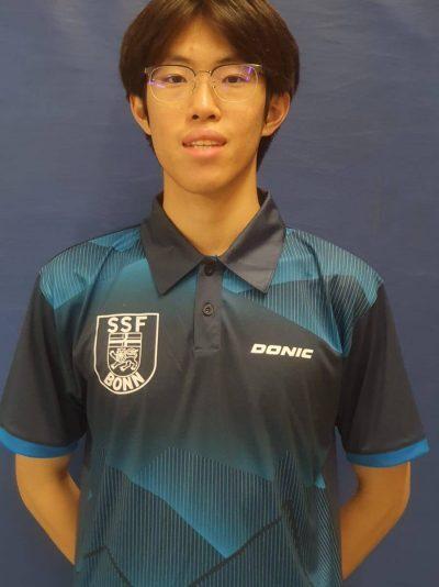 Junger Mann tr&auml;gt ein schwarzes Polo-Shirt mit blauen Akzenten und dem Logo von SSF Bonn, vor einem blauen Hintergrund.