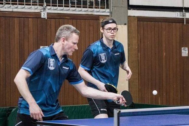Zwei Tischtennisspieler in blauen Trikots bei einem Match an einem Tischtennistisch, Ball in der Luft.