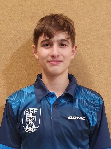 Portr&auml;t eines jungen Sportlers in blauem Tischtennisshirt mit Logo &bdquo;SSF Bonn&ldquo;, vor brauner Wand stehend.