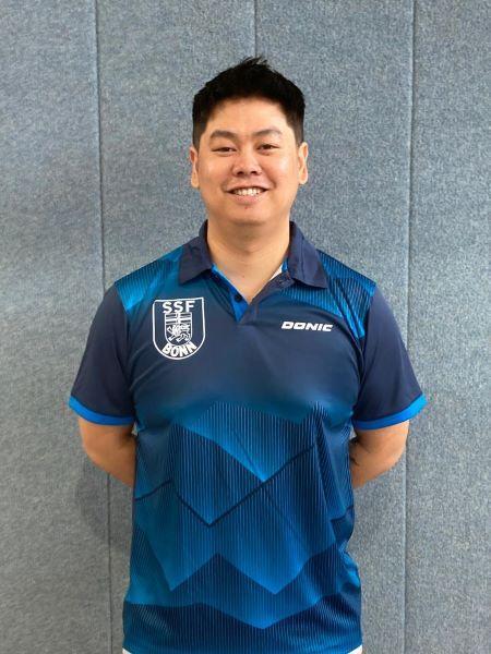 L&auml;chelnder Mann in blauem Sporttrikot mit Vereinslogo vor grauem Hintergrund.