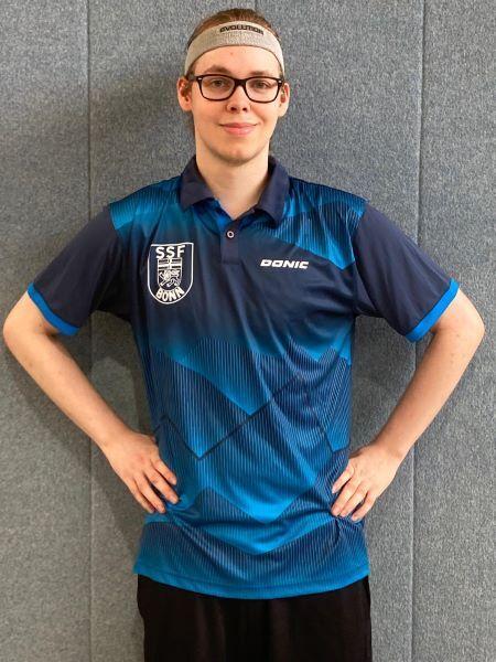 Junger Mann in einem blauen Tischtennistrikot mit Logo, steht vor einer grauen Wand und h&auml;lt seine H&auml;nde an der H&uuml;fte.